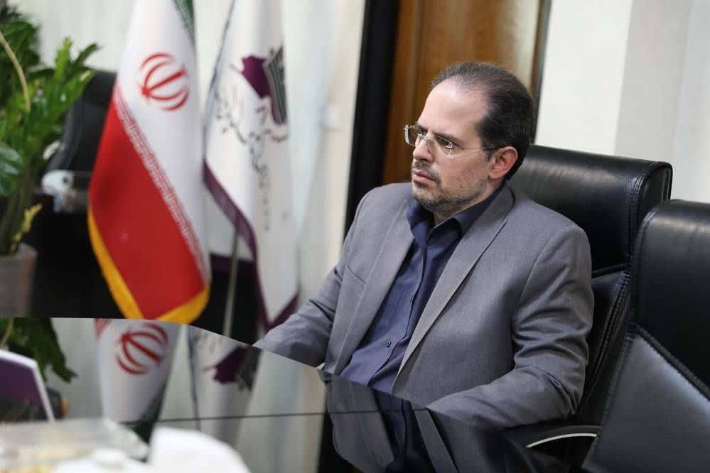 مردی با کت‌وشلوار خاکستری در حال نشستن پشت میز در یک دفتر، با پرچم ایران در پس‌زمینه